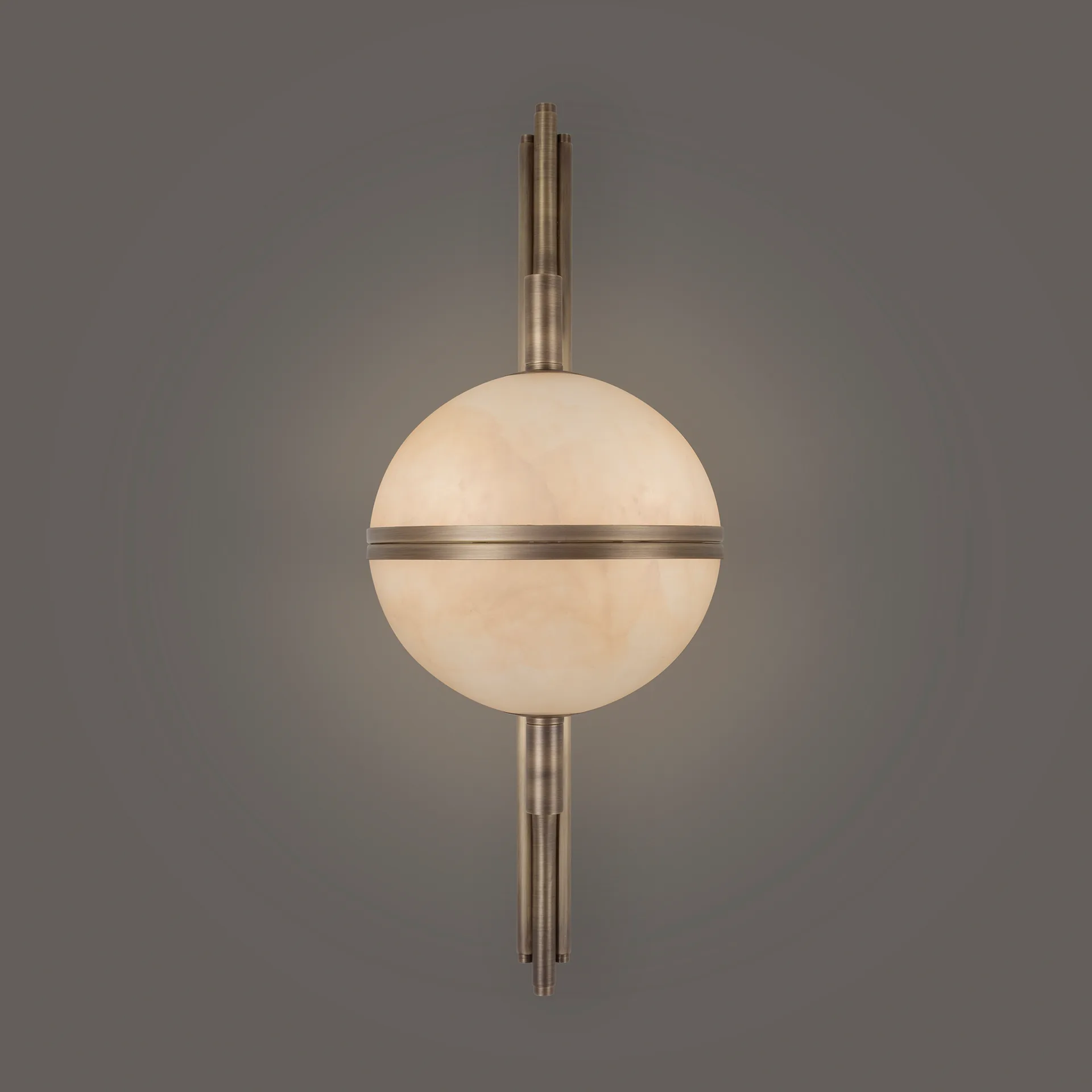 Andros 1 A detailed frontal view of the Andros wall lamp in aged brushed brass and alabaster, fully lit against a solid grey background. The central alabaster orb glows with a soft, uniform warm light, contrasting with the detailed, linear aged brass components of the stem and arms. The texture and depth of the alabaster are emphasized by the backlighting.