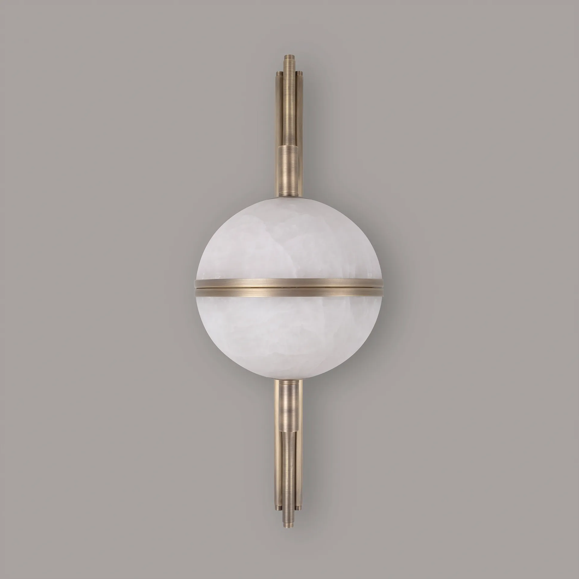 Andros 2 A clear frontal shot of the Andros wall lamp in aged brushed brass and alabaster against a solid grey background. The central orb is a full, perfect sphere of two alabaster halves separated by a horizontal brass ring. This view shows the perfect alignment of the vertical brass stem with the curved arms, and the uniform look of the natural alabaster in a passive state.