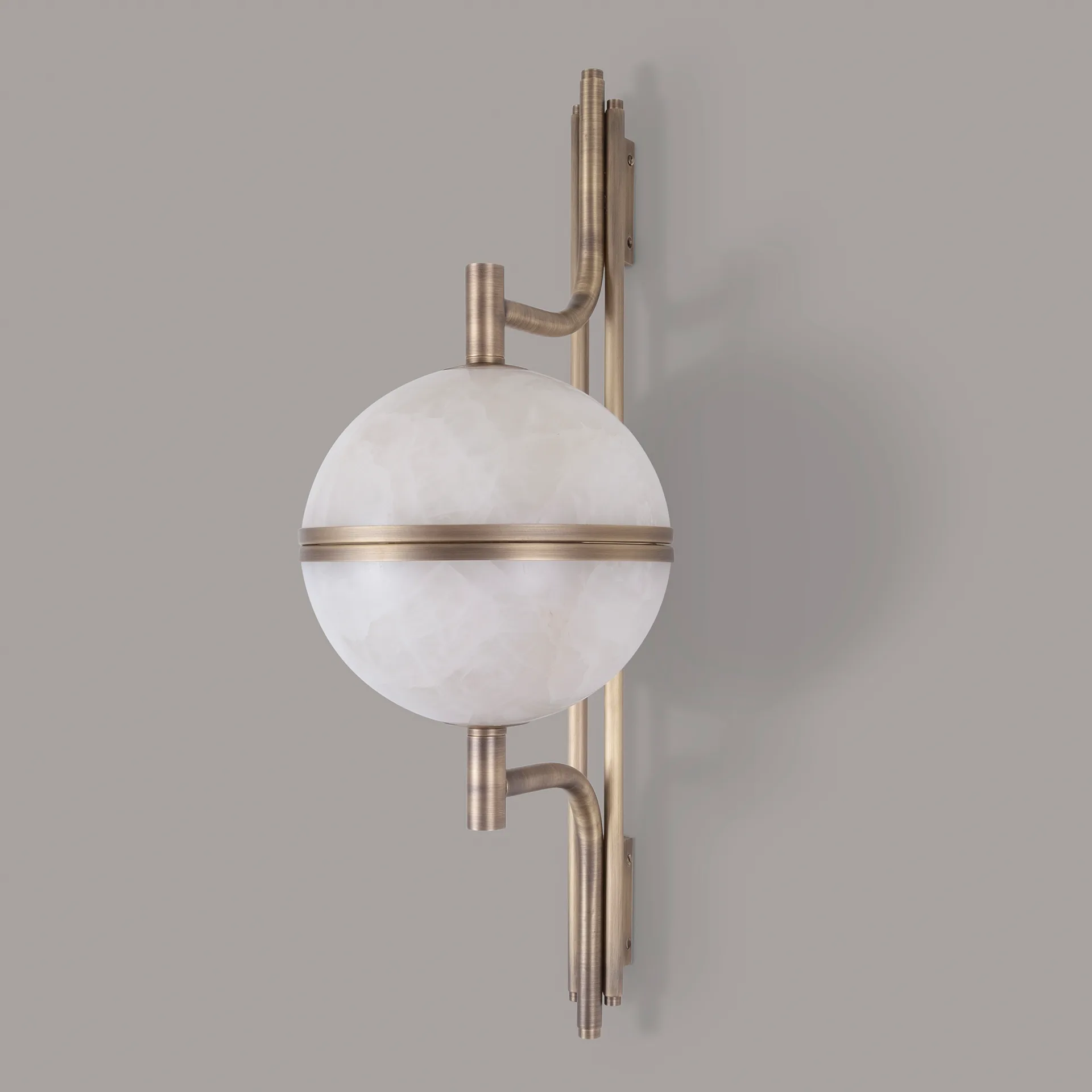 Andros 4 A clean, frontal profile view of the Andros wall lamp in aged brushed brass and alabaster, showing the full, two-hemisphere form against a grey background. This shot focuses on the symmetry of the design, with the two curved arms mirroring each other above and below the horizontal central ring. The subtle, horizontal texturing of the brushed finish is visible.