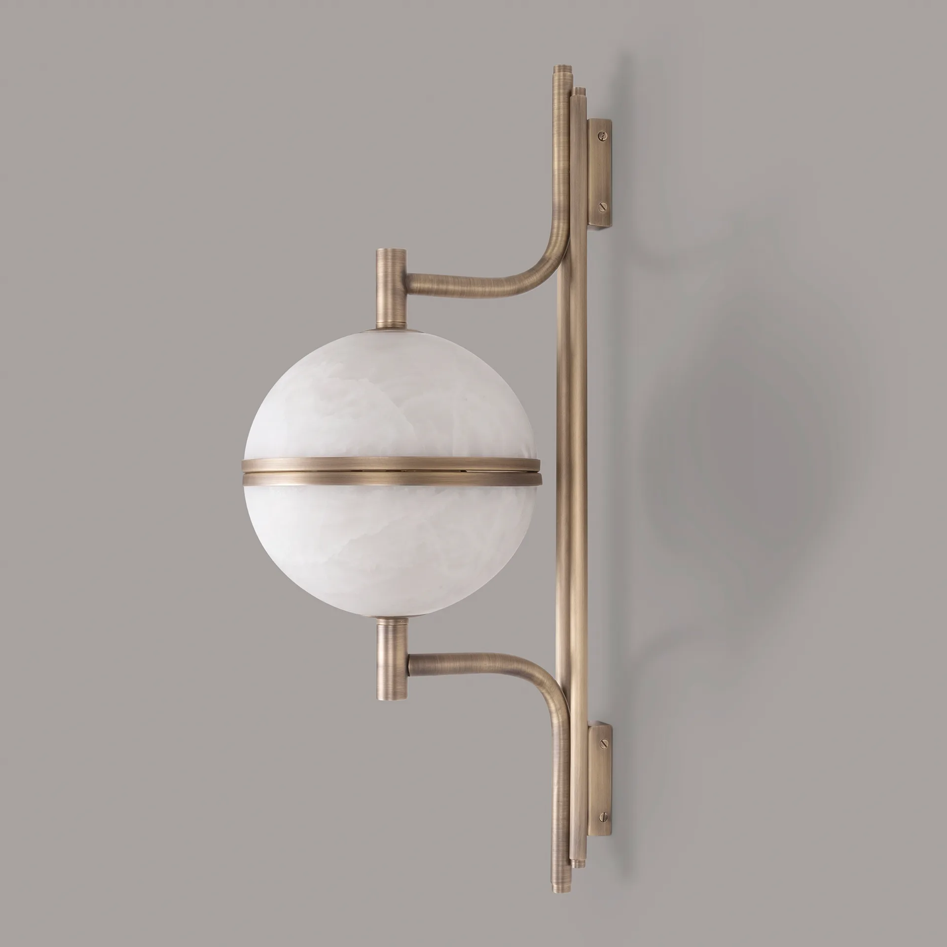 Andros 5 A clean, frontal profile view of the Andros wall lamp in aged brushed brass and alabaster, showing the full, two-hemisphere form against a grey background. This shot focuses on the symmetry of the design, with the two curved arms mirroring each other above and below the horizontal central ring. The subtle, horizontal texturing of the brushed finish is visible.