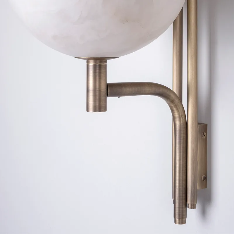 Andros Wall Lamp A angled close-up view of the Andros wall lamp in aged brushed brass and alabaster against a smooth white wall. The shot is composed to focus on the lower curved arm, its junction with the central housing, and the lower edge of the bottom translucent alabaster hemisphere.