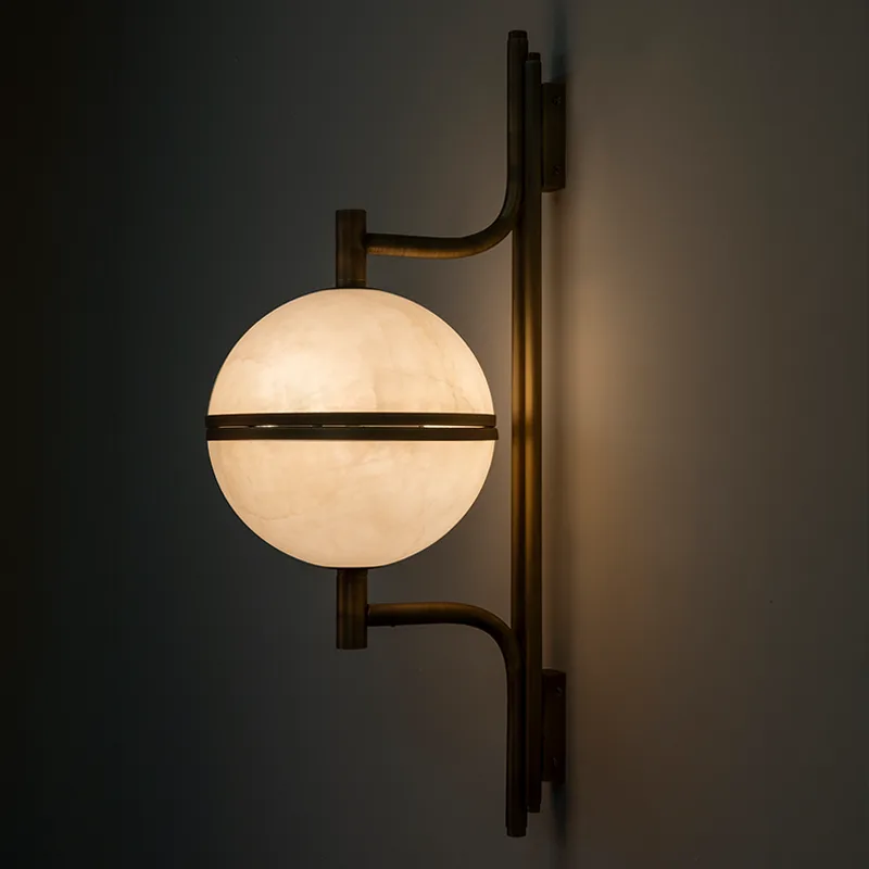Andros Wall Lamp A three-quarter angle view of the Andros wall lamp in aged brushed brass and alabaster, fully illuminated, against a smooth dark grey wall. The central alabaster globe glows with a warm light, casting shadows and reflections. This shot shows the depth of the piece, with the two curved arms holding the light source forward from the central column.