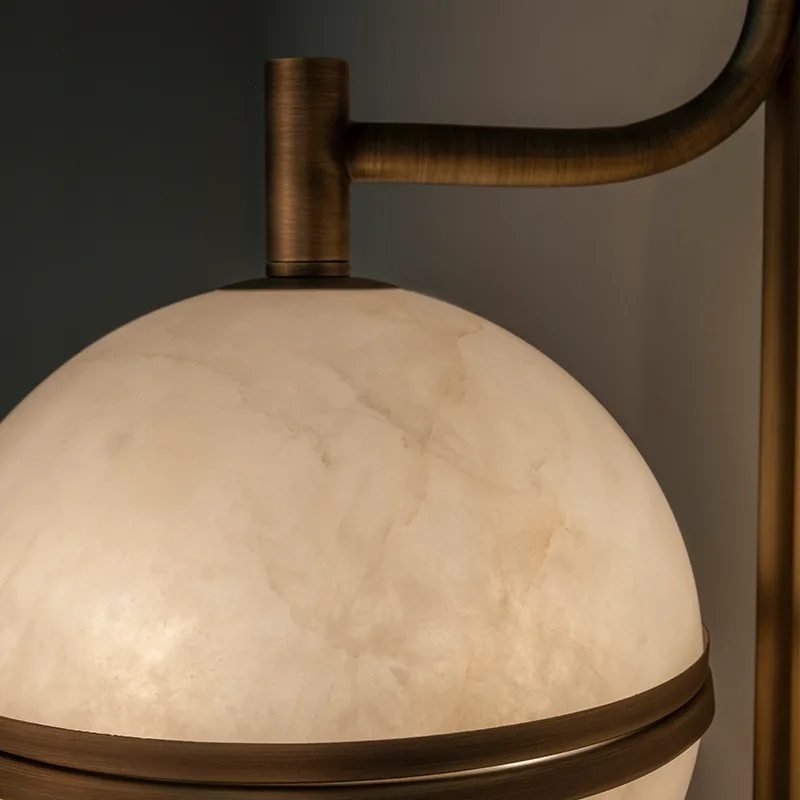 Andros Wall Lamp An close-up, focusing on the left-side joint of the Andros wall lamp in aged brushed brass and its connections to the illuminated top alabaster segment. The view highlights the material contrast and texture of the brass arm where it curves and fits into the central brass ring.