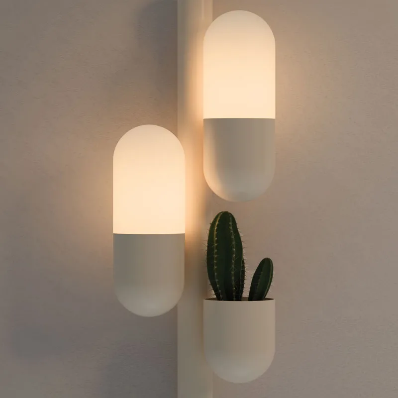 Cactus wall lamp detail with its two lights turned on, emitting a warm glow, lacquered in cream color with white glass shades. The piece also features a succulent plant.