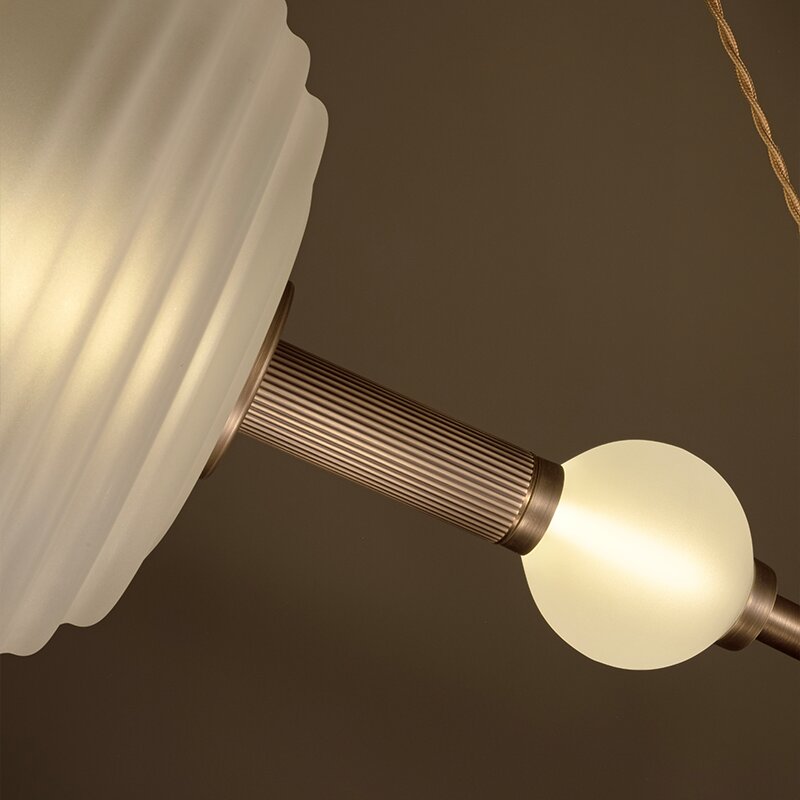 Saturn Suspension Lamp Detailed close-up showing the craftsmanship of the Saturn suspension lamp. It highlights a glowing, small frosted glass bulb attached to a fluted antique brass metal rod, which extends from a larger frosted glass ribbed globe. The warm, soft glow illuminates the detailed texture of the metalwork against a moody brown background.