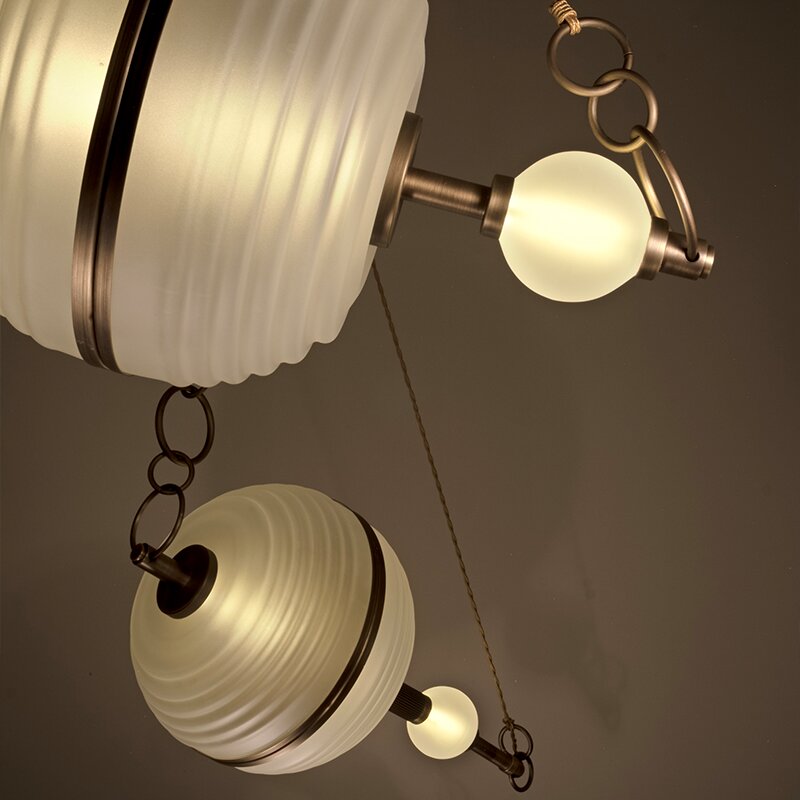Saturn Suspension Lamp A low-angle view showcasing two Saturn suspension lamps hanging at different heights. The modern luxury lighting fixtures feature ribbed frosted glass spheres resembling planets with central brass bands, paired with smaller glowing frosted glass bulbs and hanging from matching antique brass link chains and slender cords against a dark ambient backdrop.