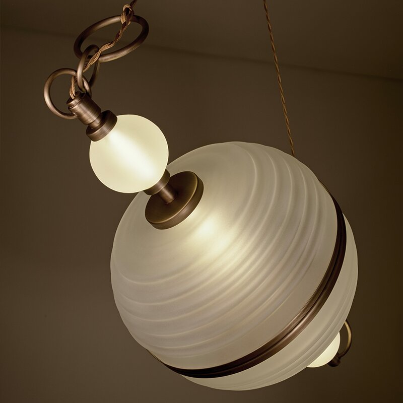 Saturn Suspension Lamp Close-up of the Saturn suspension lamp by Creativemary against a dark warm background. The lighting fixture features a large, spherical, frosted ribbed glass globe reminiscent of a planet, encircled by an antique brass metal ring. A smaller, illuminated spherical frosted glass bulb is attached to the brass fixture above, connected to a textured metal chain and a thin power cord.