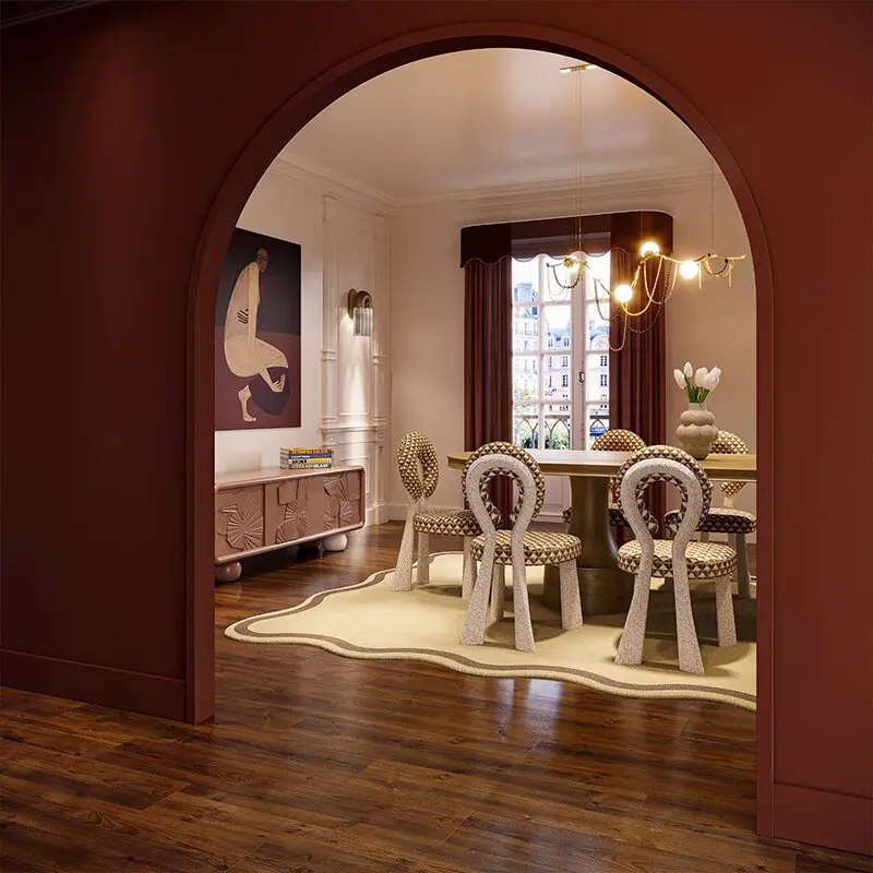A view through a deep terracotta-red archway into a sophisticated, eclectic dining room. The space features a set of mary dining chairs with unique looped backrests and patterned upholstery surrounding a buck oval dining table. Above the table hangs the sculptural, branching berlin suspension lamp, while a silo wall lamp provides soft accent lighting next to a modern figurative painting. To the left, a textured mon ooh sideboard in a dusty rose finish sits against a white paneled wall, all tied together by a wavy-edged cream rug on dark wood floors.